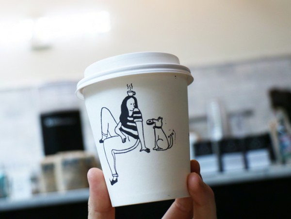 Hand holding up paper coffee with a stamped illustration of girl sitting beside dog