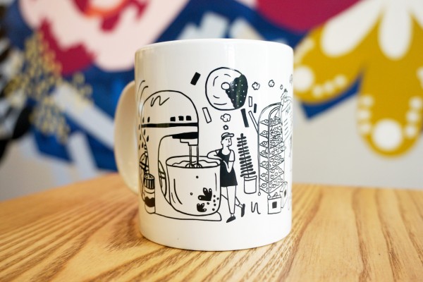 White ceramic mug on wooden table with colorful mural in background. Mug illustration includes oversized kitchen items and bakers. 
