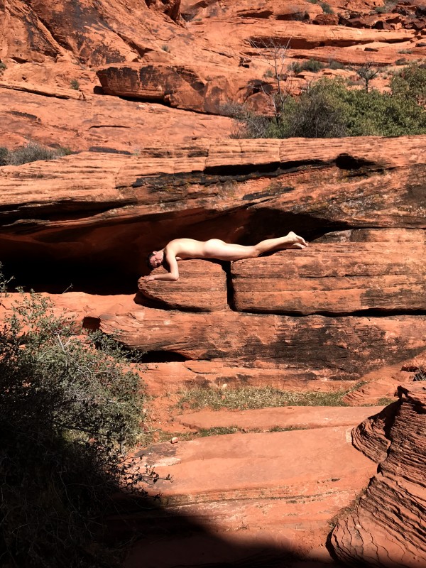 Photograph of red rocks. Male model lays horizontal and nude in crevice of rock. 