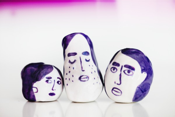 Three circular white porcelain rattles painted with purple faces on white background. 