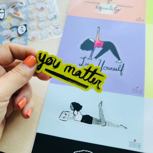 Hand with orange finger nails holding sticker that reads "You matter" with other stickers in background. 