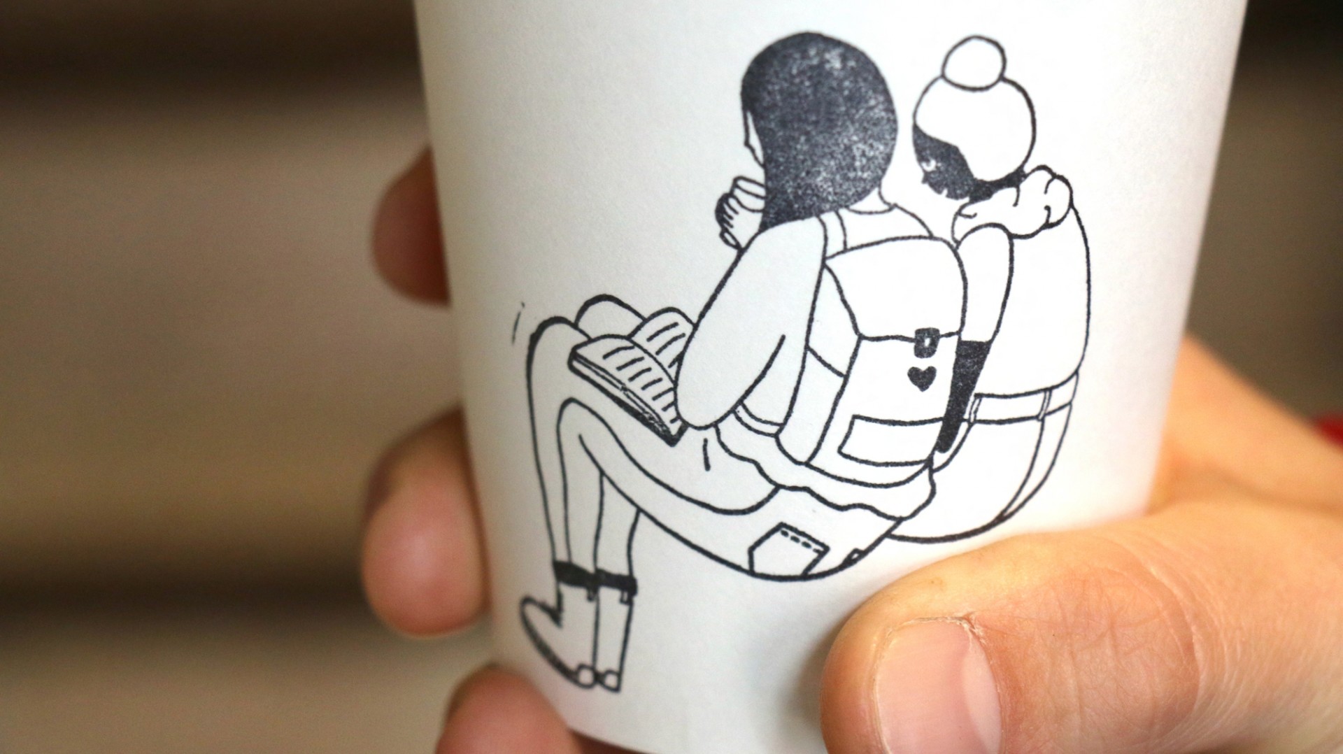 Hand holding coffee up with a stamped illustration of two girls sitting together