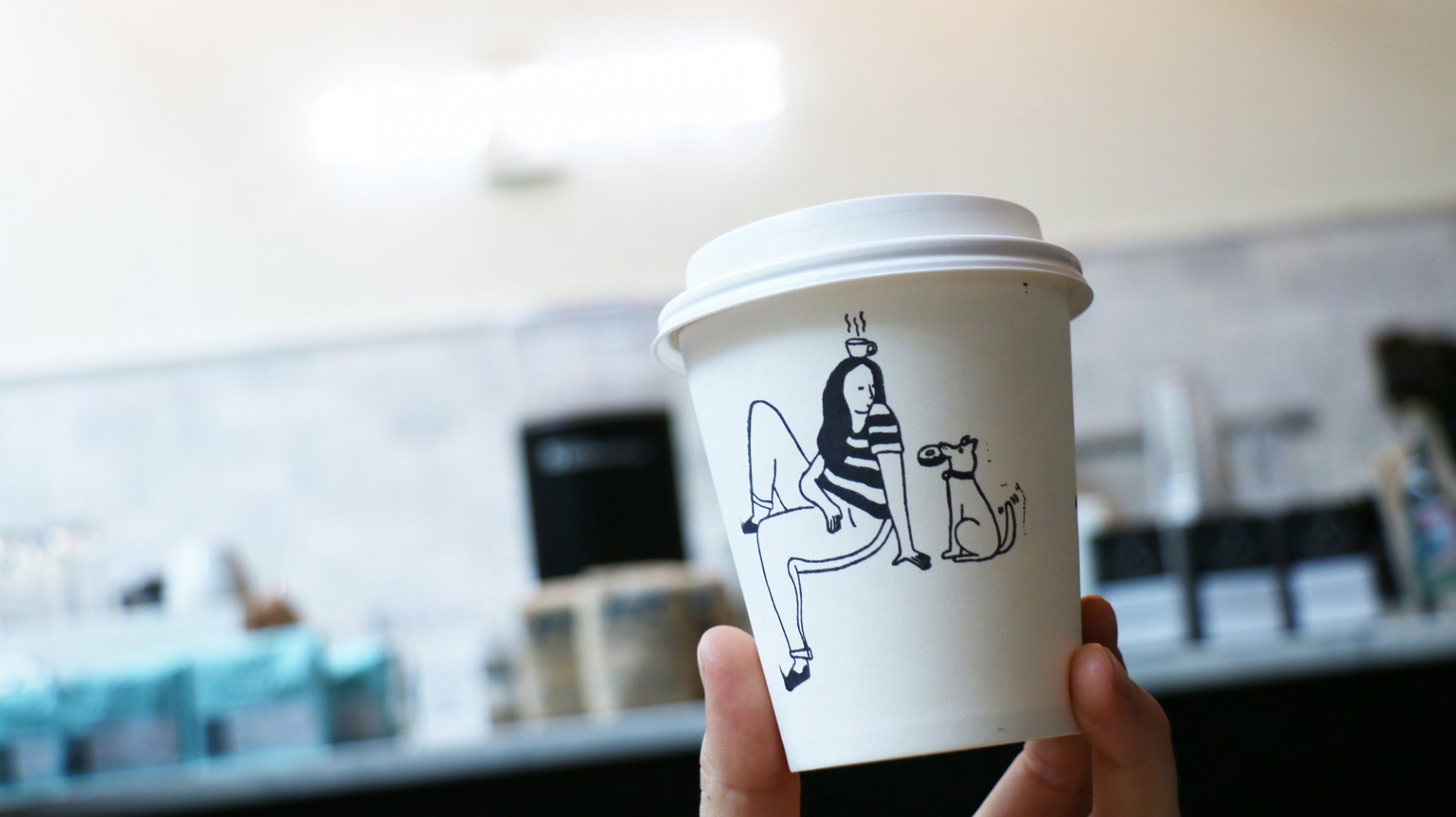Hand holding up paper coffee with a stamped illustration of girl sitting beside dog