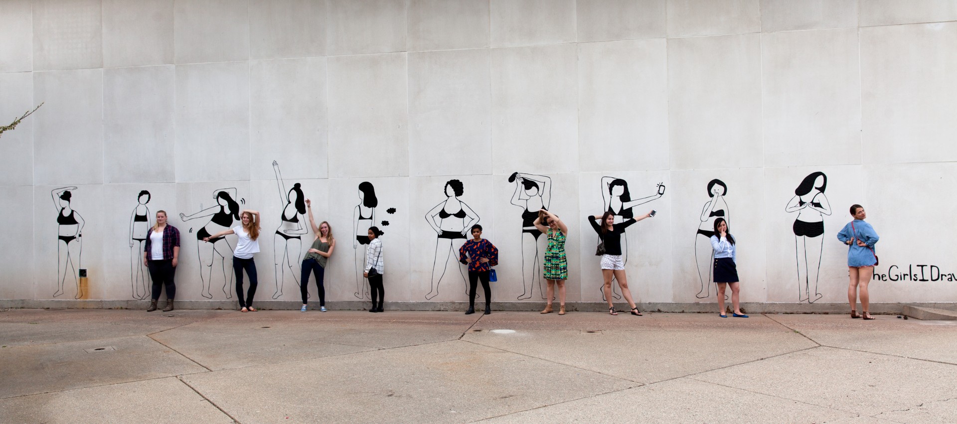 Photograph of wall with mural. Mural consists of 10 women in undergarments in various poses. Women are all rendered in black lines made out of duct tape. People are posed in front of each figure in mural mimicking the pose.