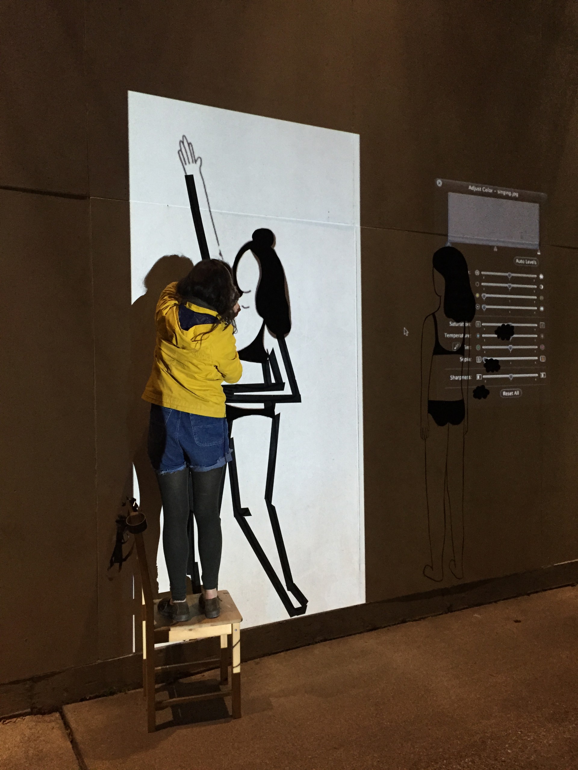 Image of artist making duct tape mural at night. She is standing on chair cast in light from projected image onto wall.