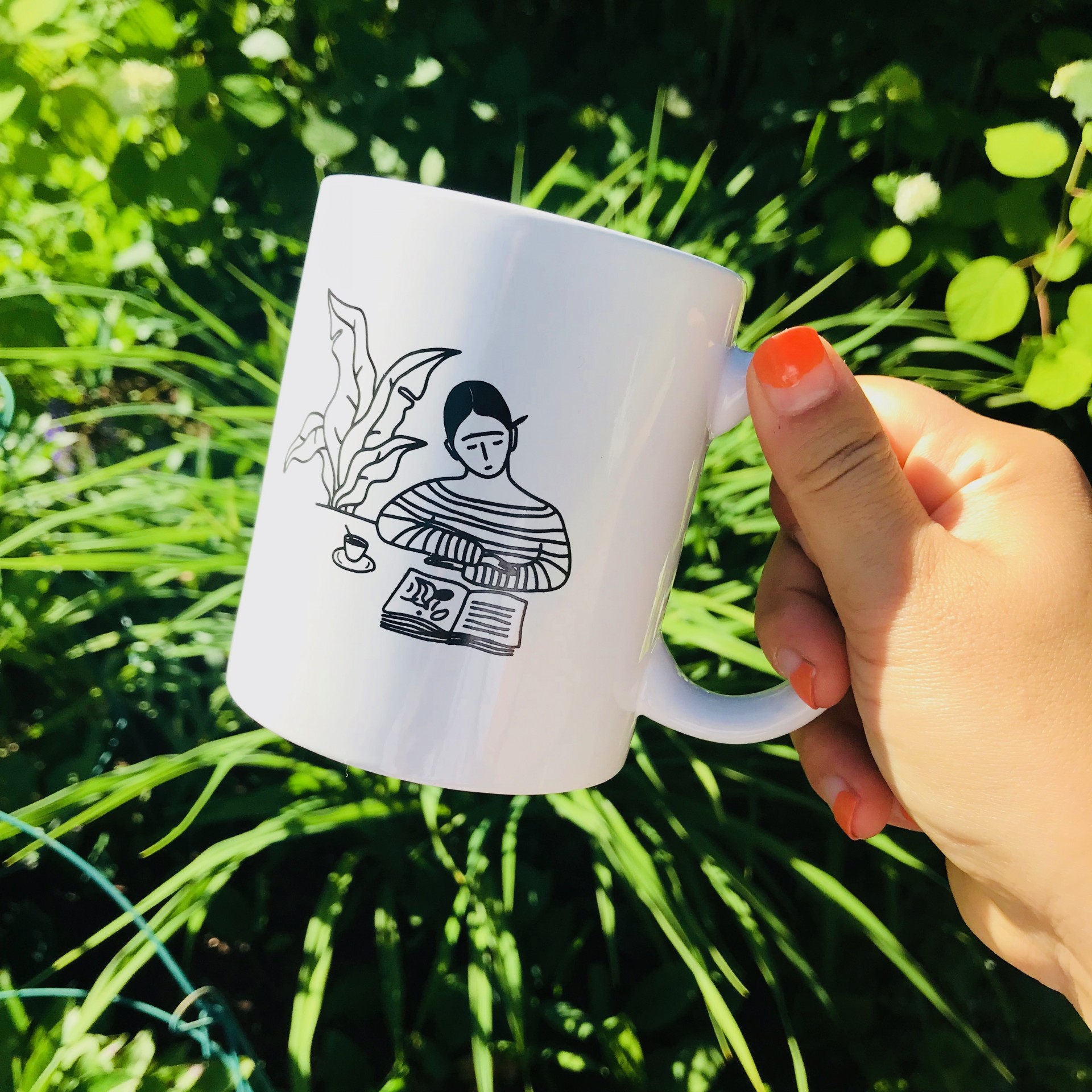Hand holding white ceramic mug. Simple black drawing on mug features girl in striped shirt reading at table with plant to her left. 