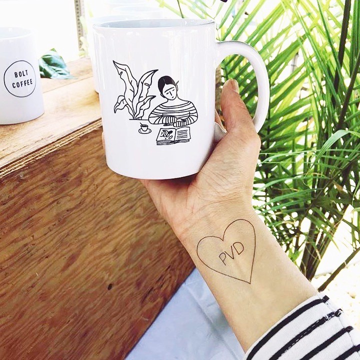 Hand holding white ceramic mug. Simple black drawing on mug features girl in striped shirt reading at table with plant to her left. Hand holding mug has a tattoo of a heart with the text "PVD" inside. 