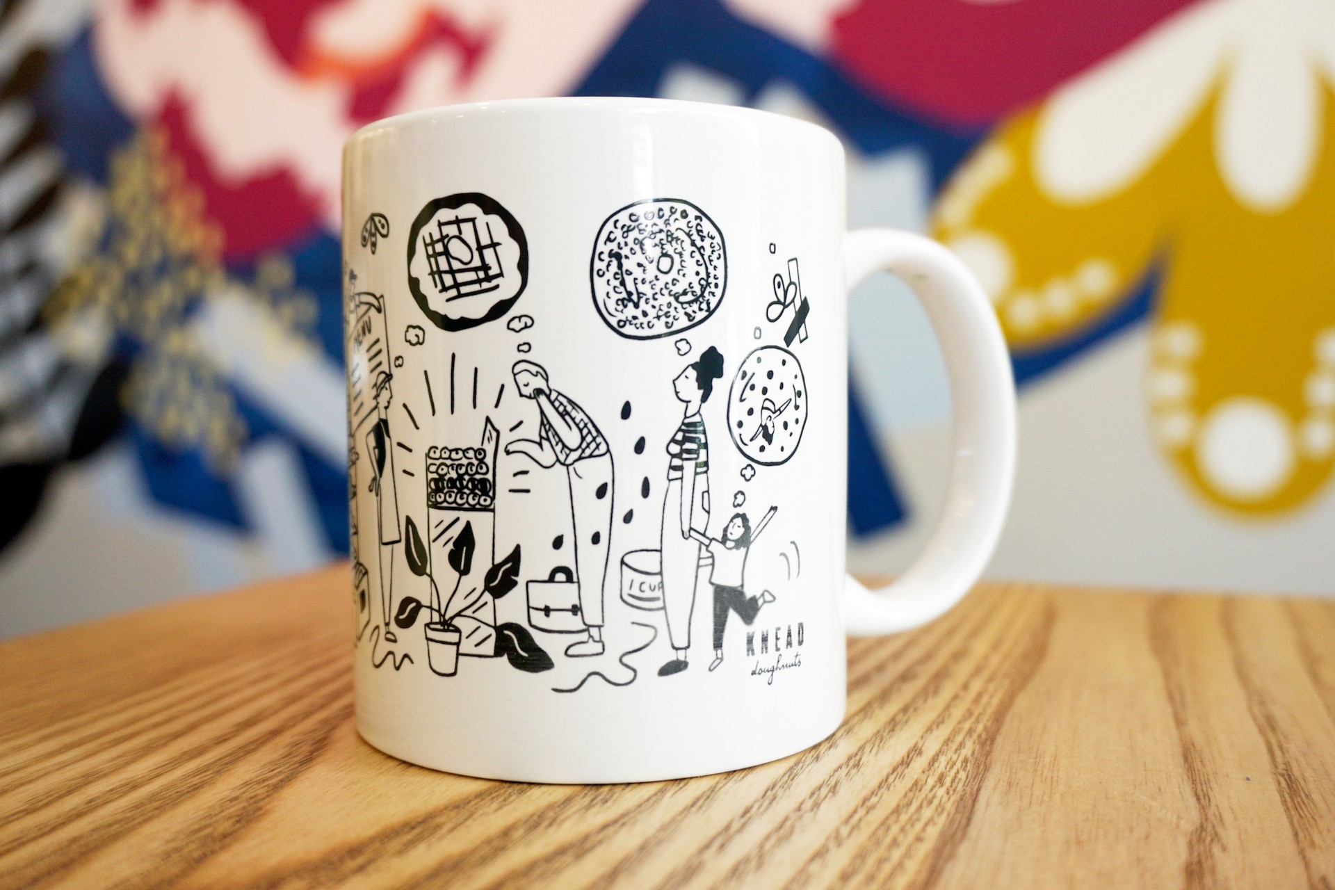 White ceramic mug on wooden table with colorful mural in background. Mug illustration includes oversized kitchen items, donuts, bakers and Knead customers.. 