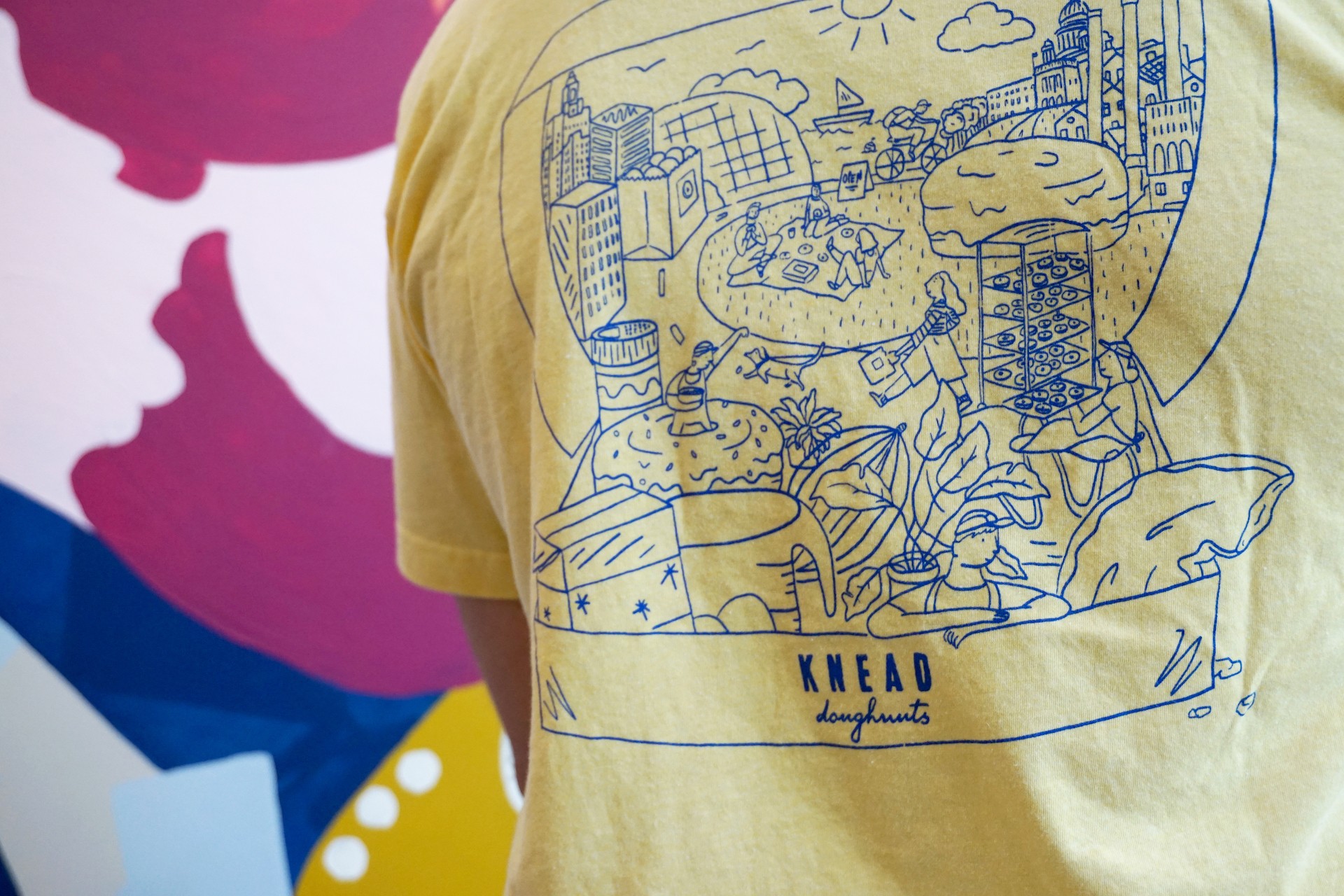 Detail of full back design of yellow t shirt. Text reads "Knead Doughnuts" Illustration shows oversized baking items, Knead employees and plants all inside Knead box of doughnuts. 