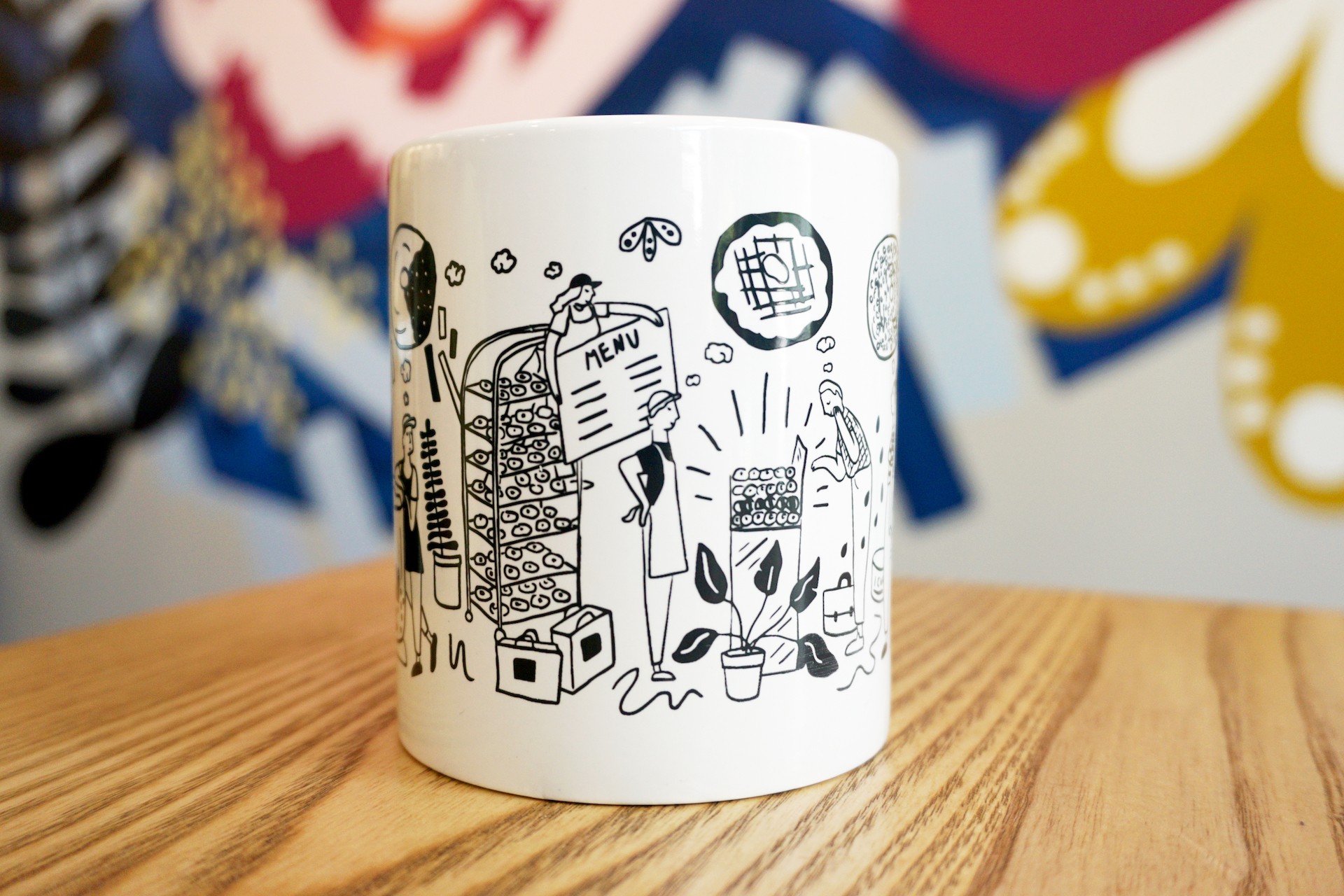 White ceramic mug on wooden table with colorful mural in background. Mug illustration includes oversized kitchen items, donuts and bakers. 