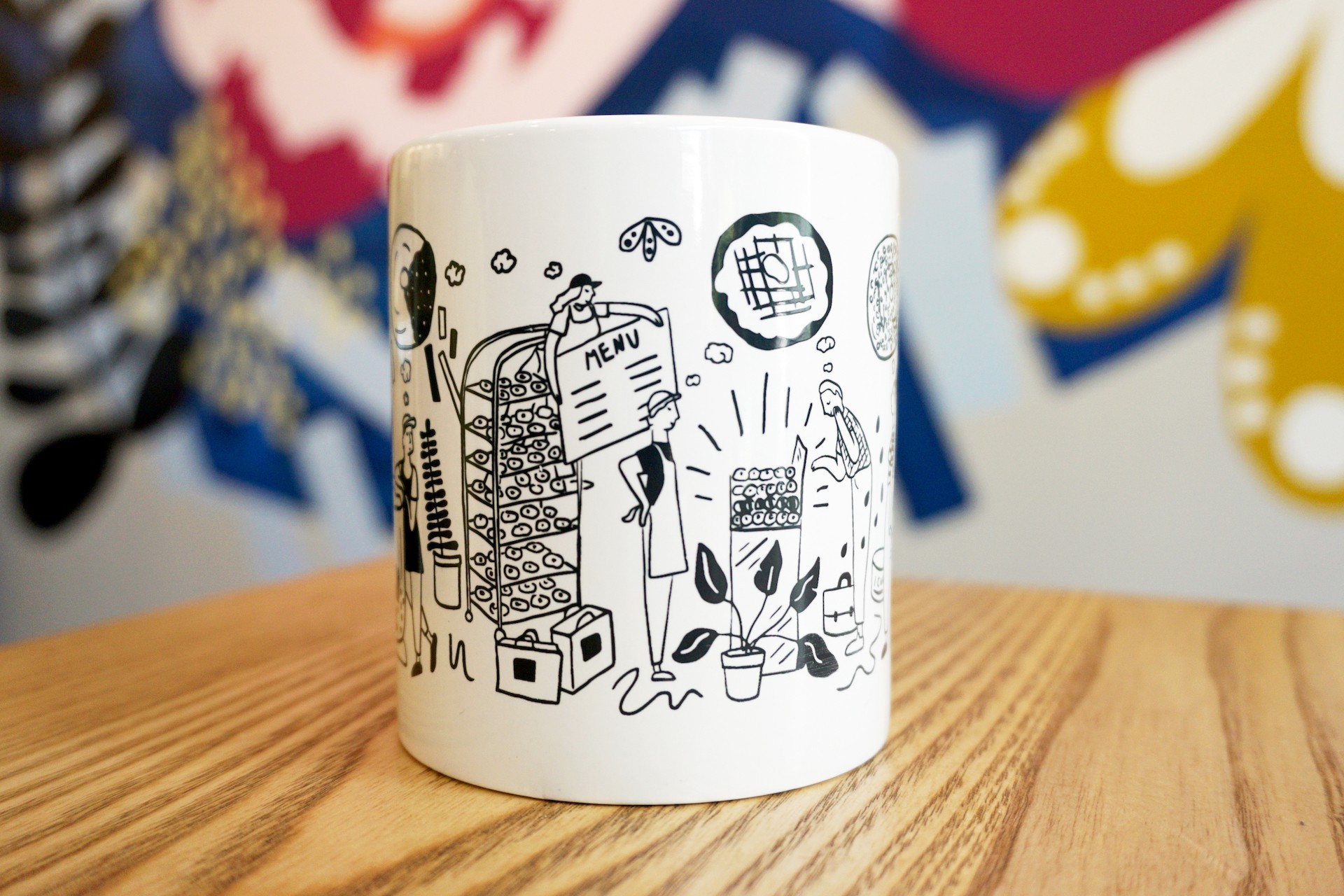White ceramic mug on wooden table with colorful mural in background. Mug illustration includes oversized kitchen items, donuts and bakers. 