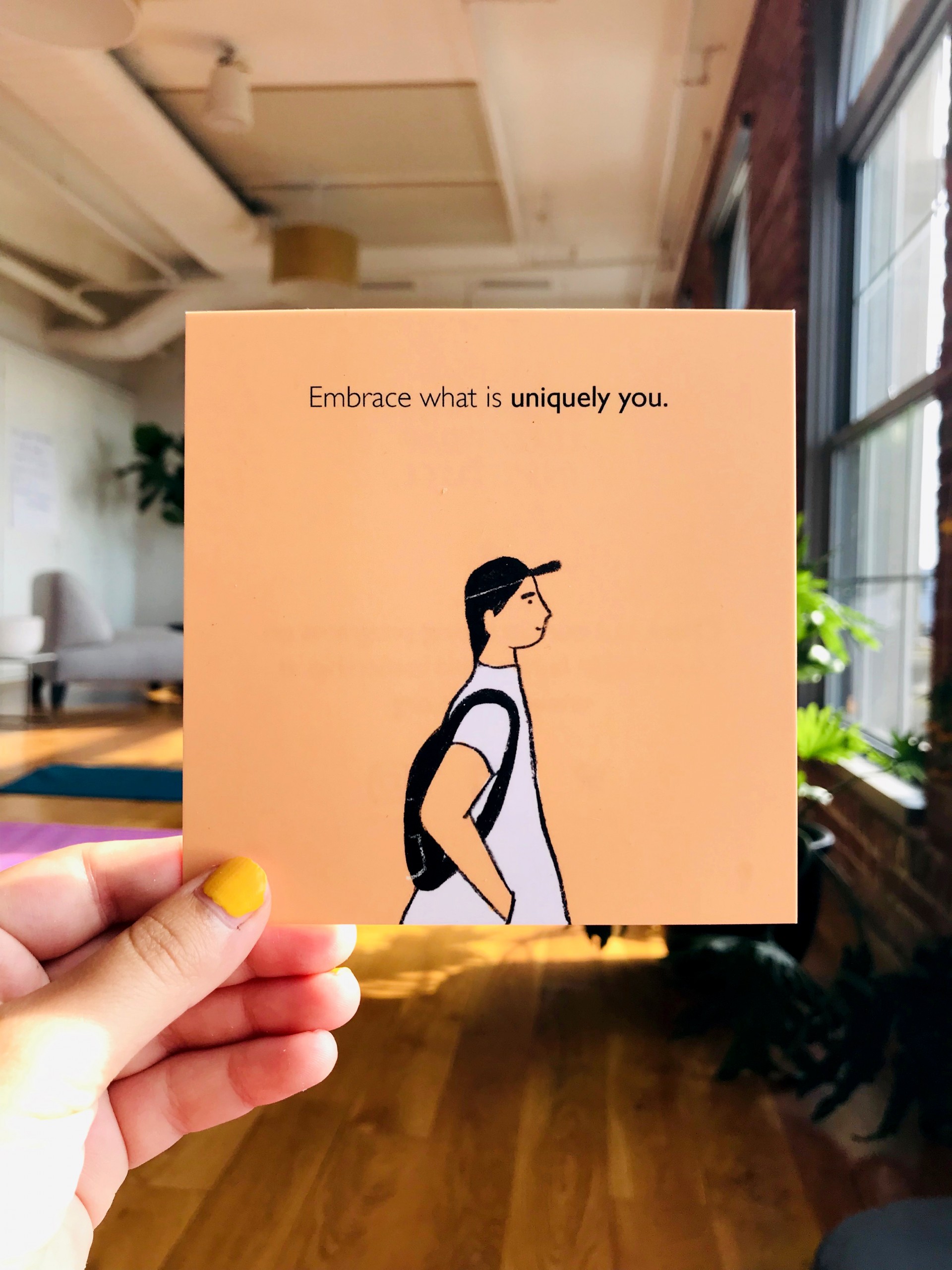 Hand holding orange postcard with figure facing side wearing backpack, hat and white dress. Text reads, "Embrace what is uniquely you"