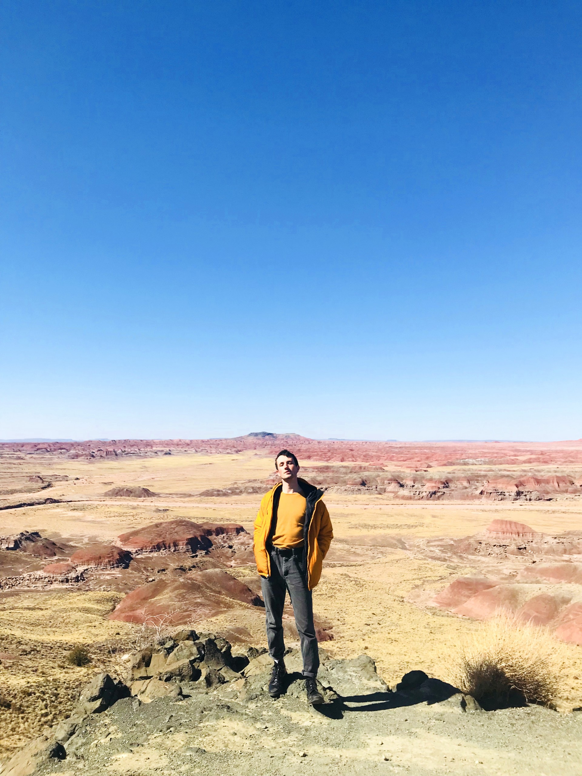 Flat southwestern yellow landscape. Male model in central foreground of image wearing yellow shirt and pants. In contrast to clear blue sky.