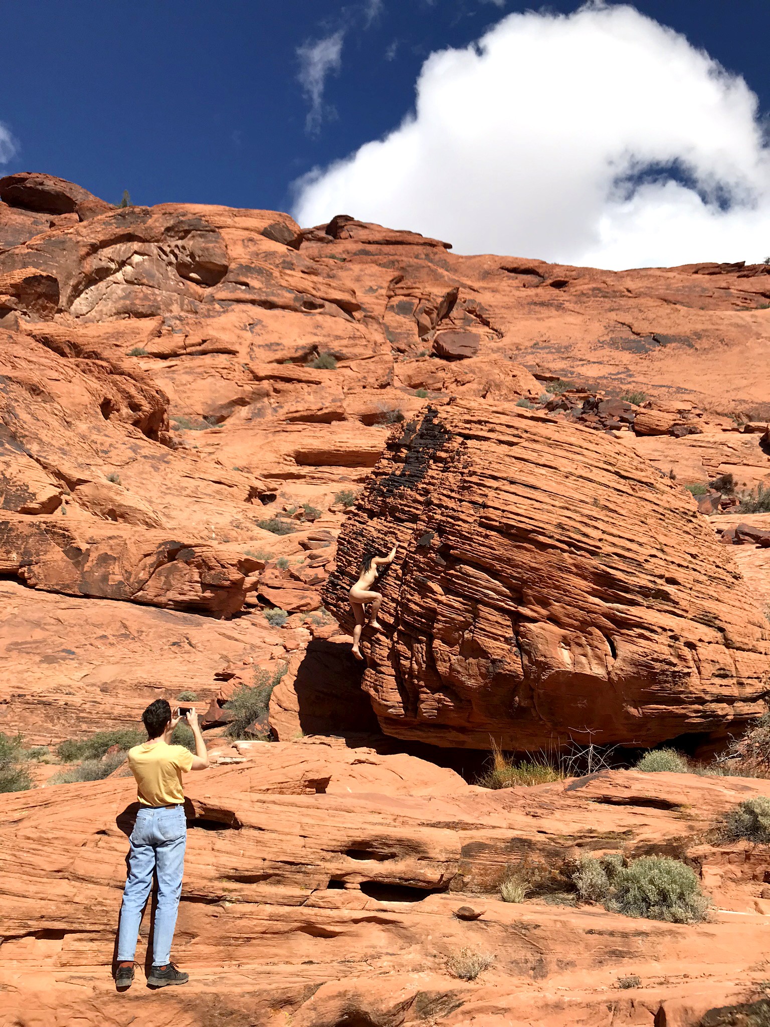Photograph of Nevada canyons. 3/4 of image are bright red rocks. Figure in bottom left corner has back towards viewer and is taking picture of nude woman climbing boulder in right side of photograph. Sky is blue with clouds.