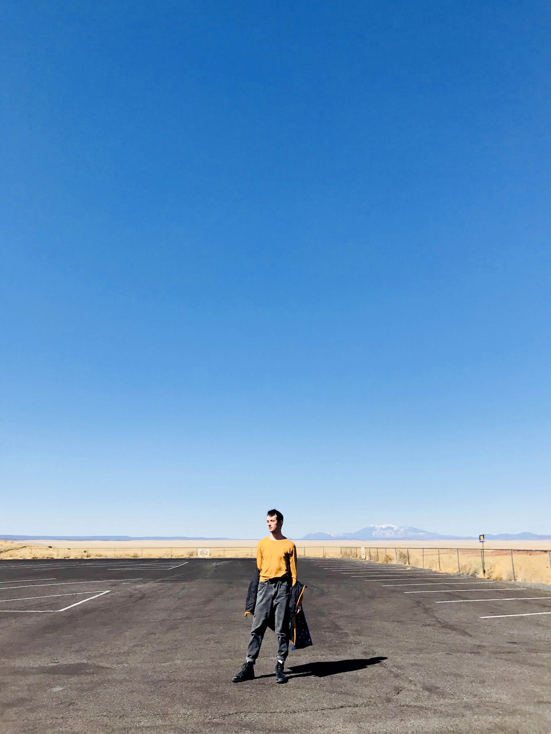 Male model standing in empty parking lot. 3/4 photograph is blue sky. Model has jacket half hanging off arms. 