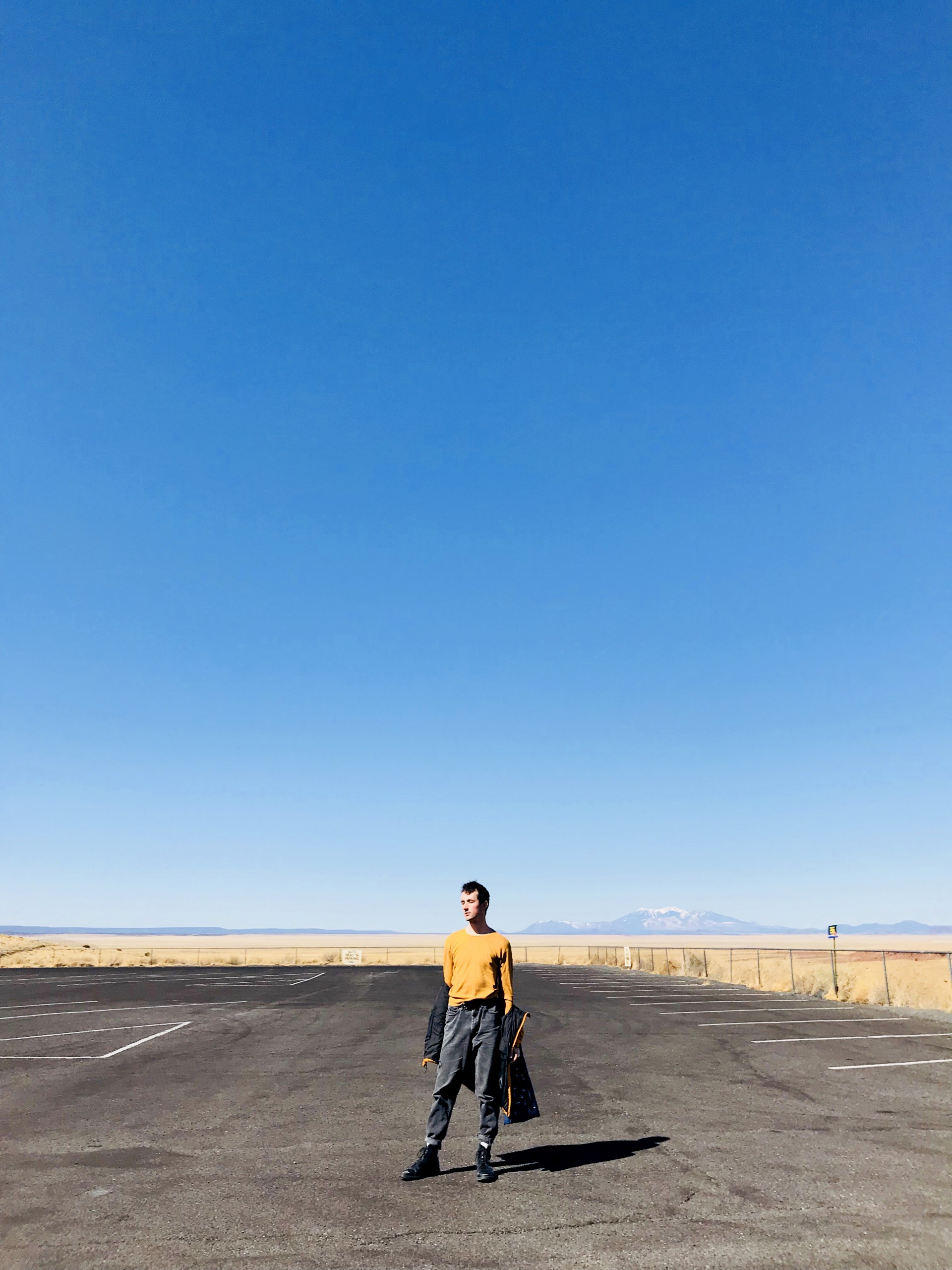 Male model standing in empty parking lot. 3/4 photograph is blue sky. Model has jacket half hanging off arms.
