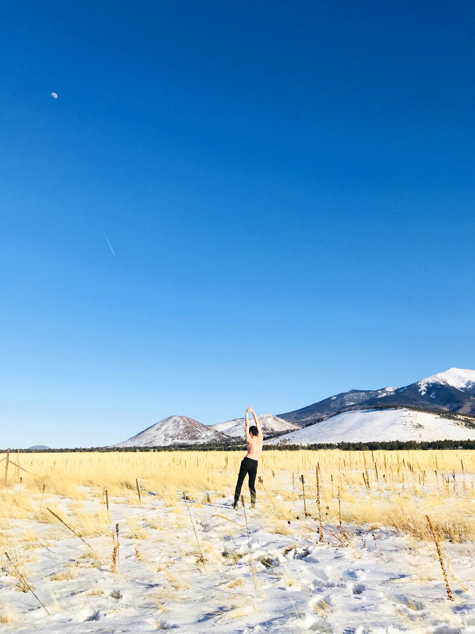 Photograph of snowy wheat field. Male model stands with back towards viewer in center of image. Body is slightly curved. Sky is blue.