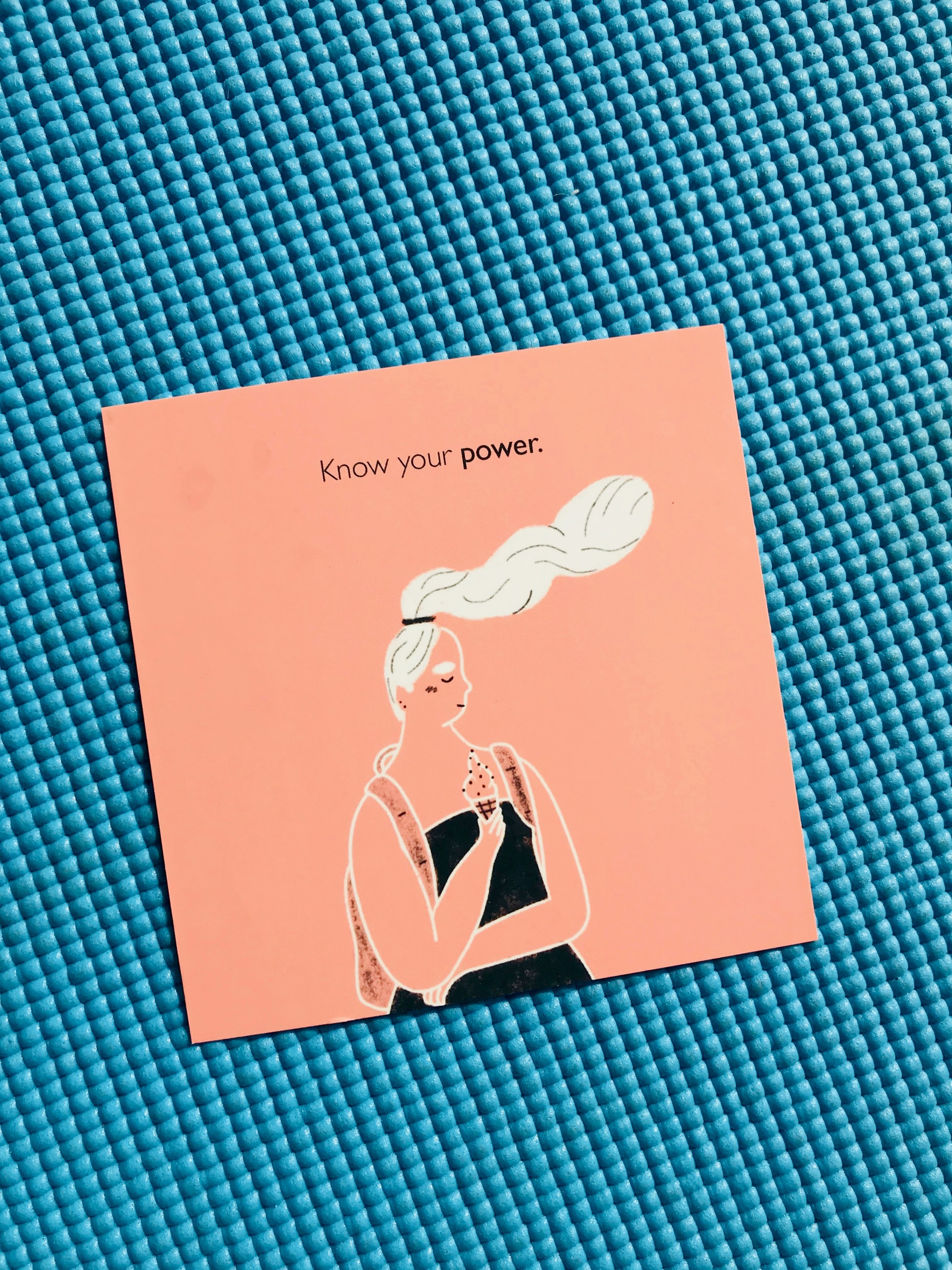 Pink square postcard against blue yoga mat. Figure on postcard is of girl with hair blowing in wind eating ice cream. Text reads, "Know Your Power"