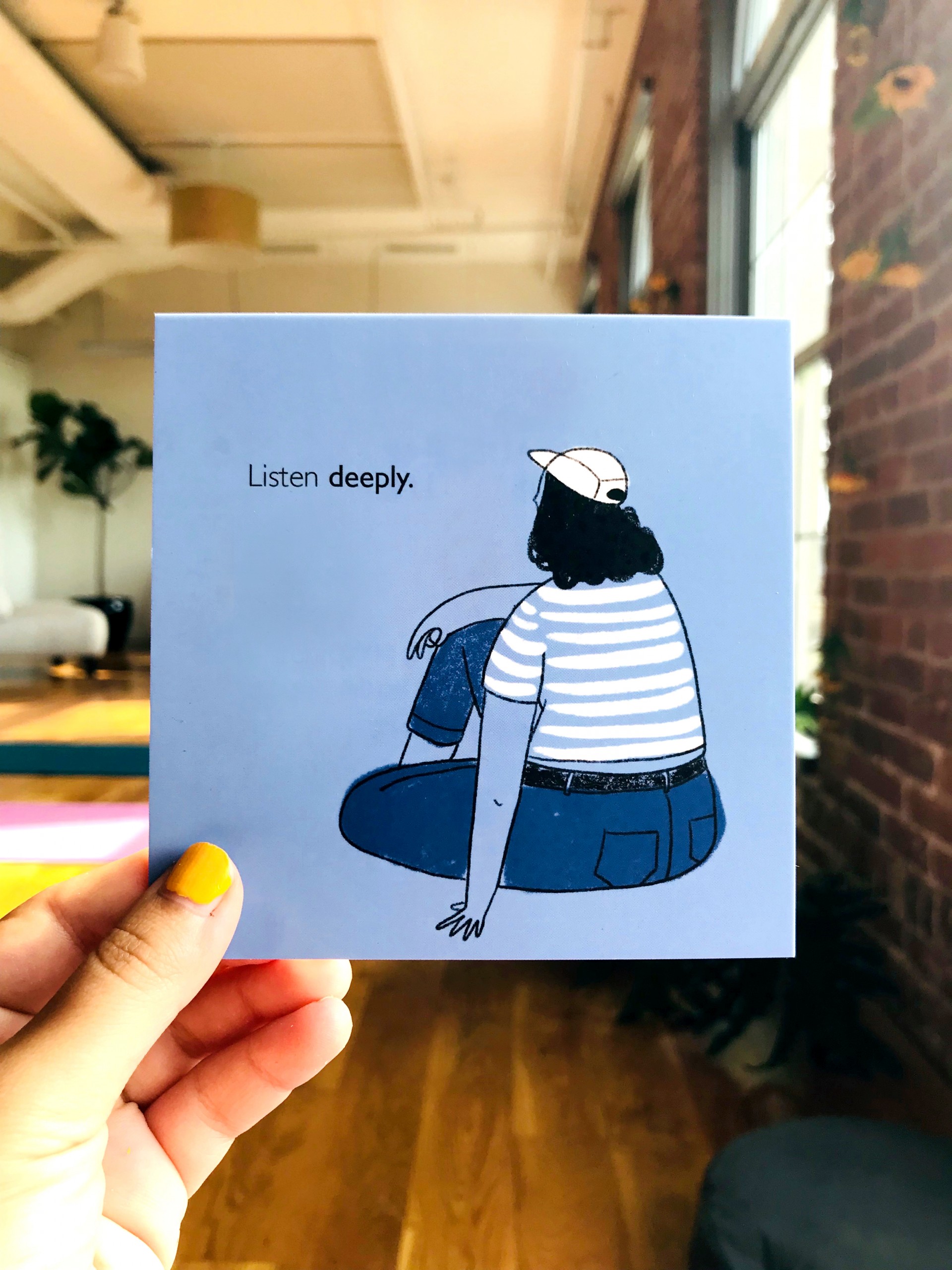 Hand holding blue postcard with figure whose back is towards viewer wearing hat and striped shirt. Text reads, "Listen Deeply"