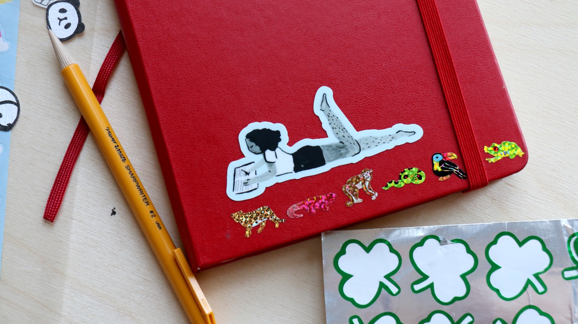 Red notebook with a blue sticker placed on top of it. The sticker is a girl laying on her stomach journaling.