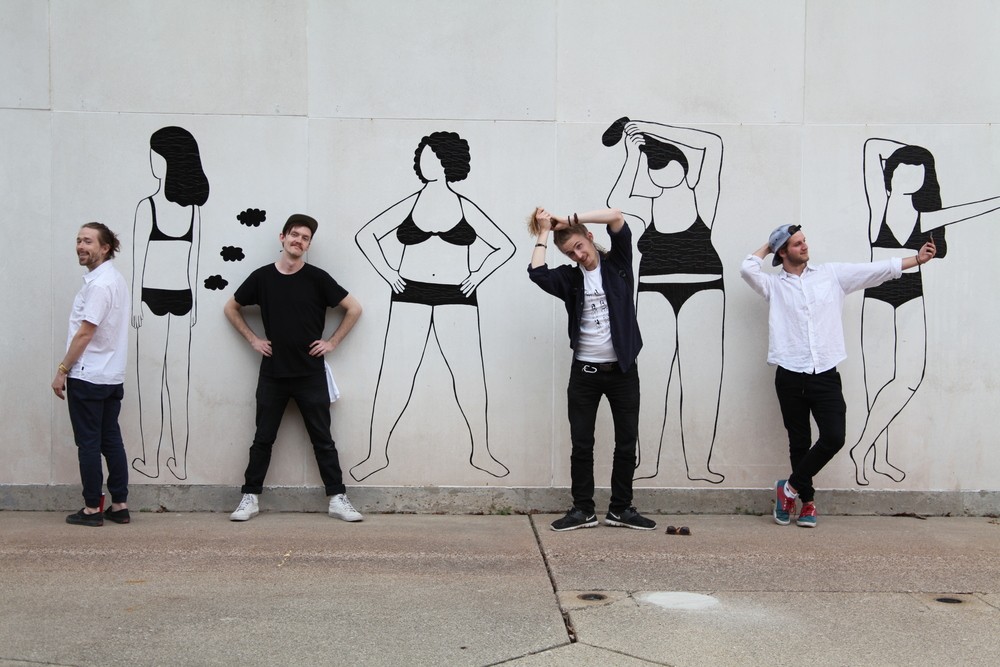 Photograph of wall with mural. Photograph shows 4 women in undergarments in various poses. Women are all rendered in black lines. People are posed in front of each figure in mural mimicking the pose.