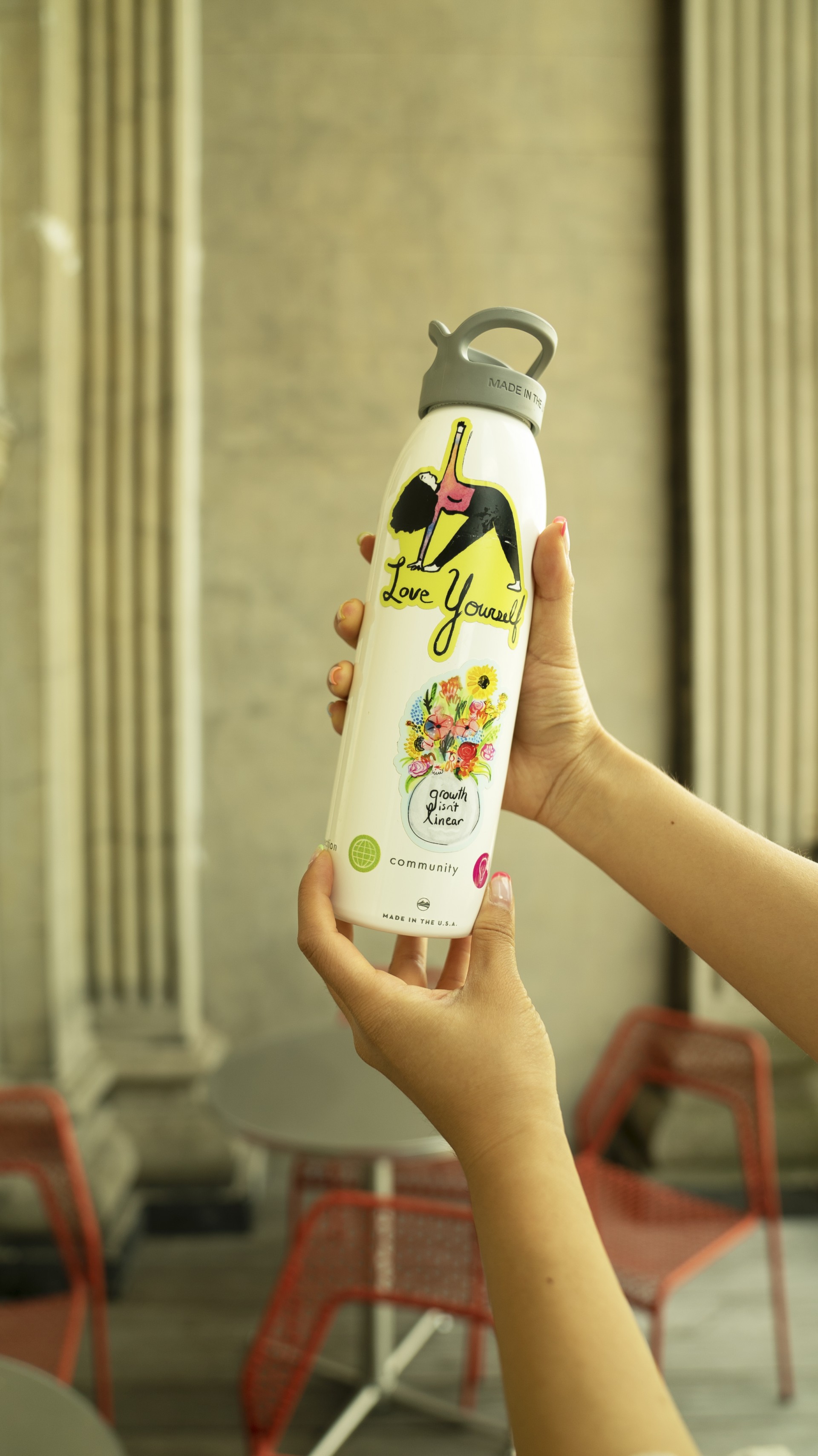 Hands holding water bottle with various stickers placed on top. One sticker reads "Love Yourself." The other reads "Growth isn't linear"