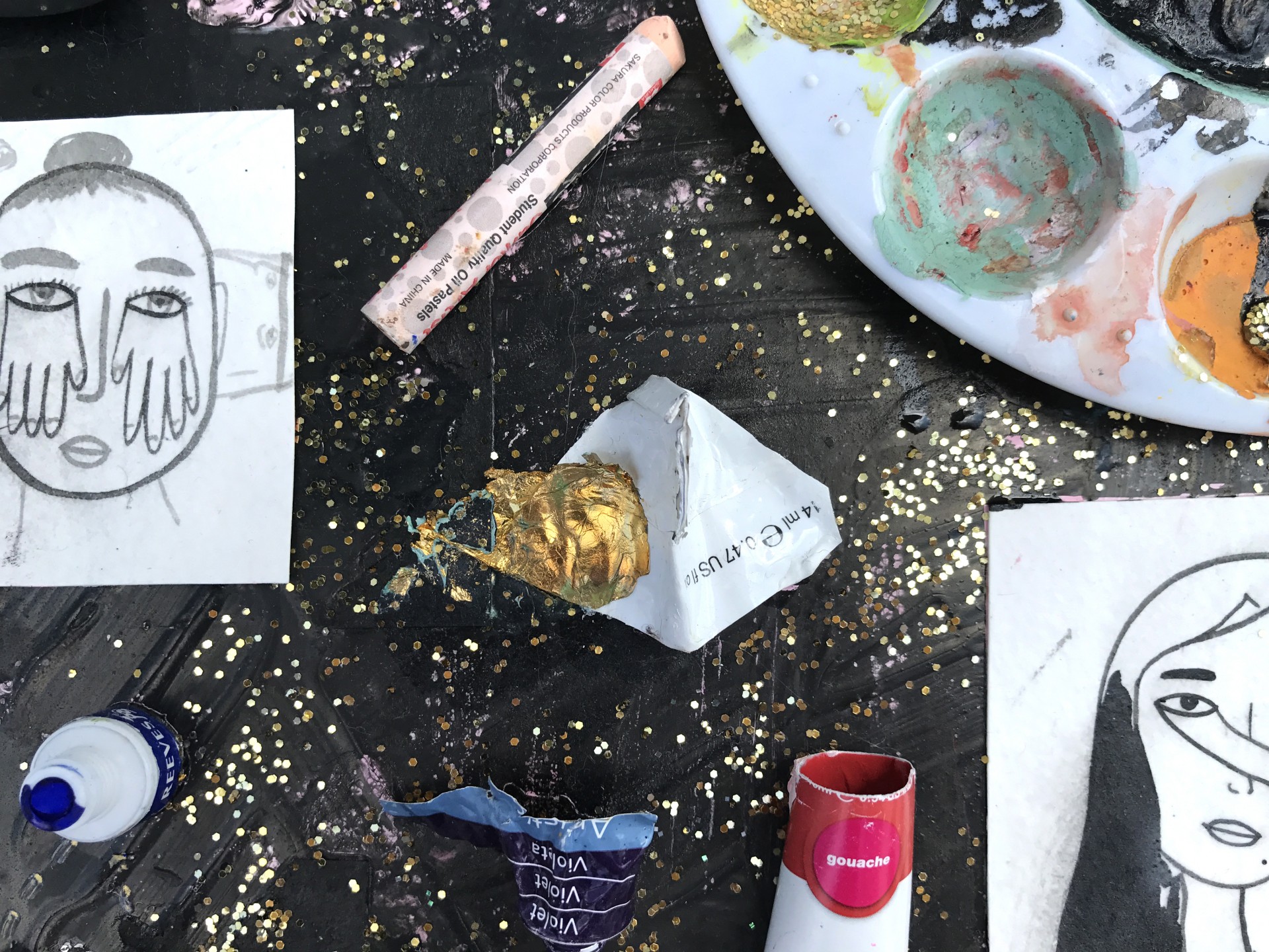 Up close view of painting. Background is black with gold glitter. Old paint tubes, paint palette and drawing of girl with hands coming from her eyes glued on top. 