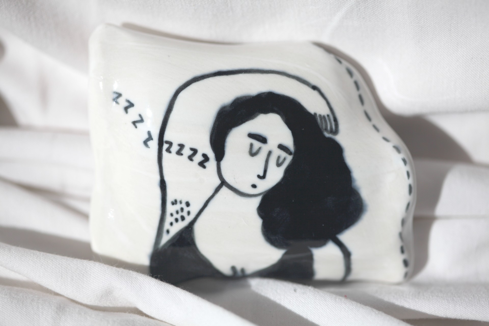 One porcelain pillow painted with a girl sleeping on it photographed against white sheets. 