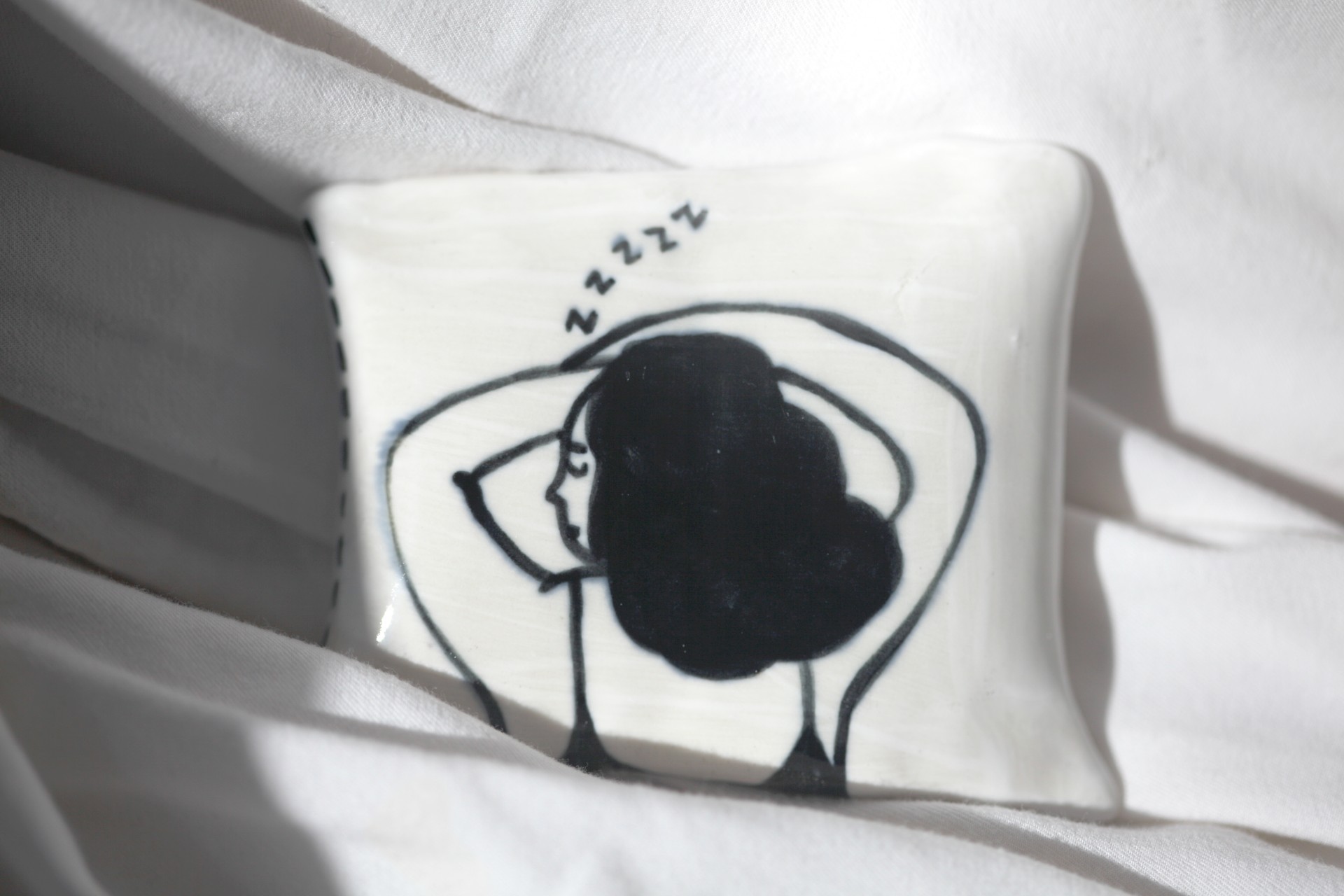 One porcelain pillow painted with a girl sleeping on it photographed against white sheets. 
