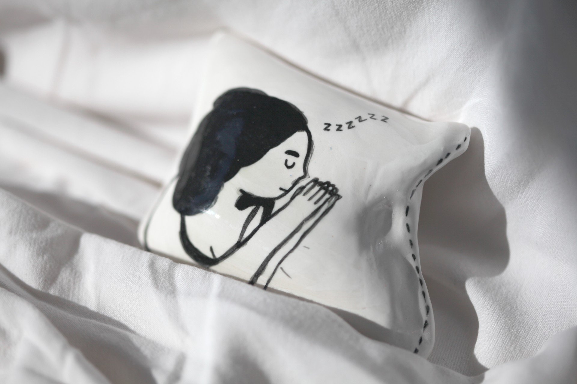 One porcelain pillow painted with a girl sleeping on it photographed against white sheets. 