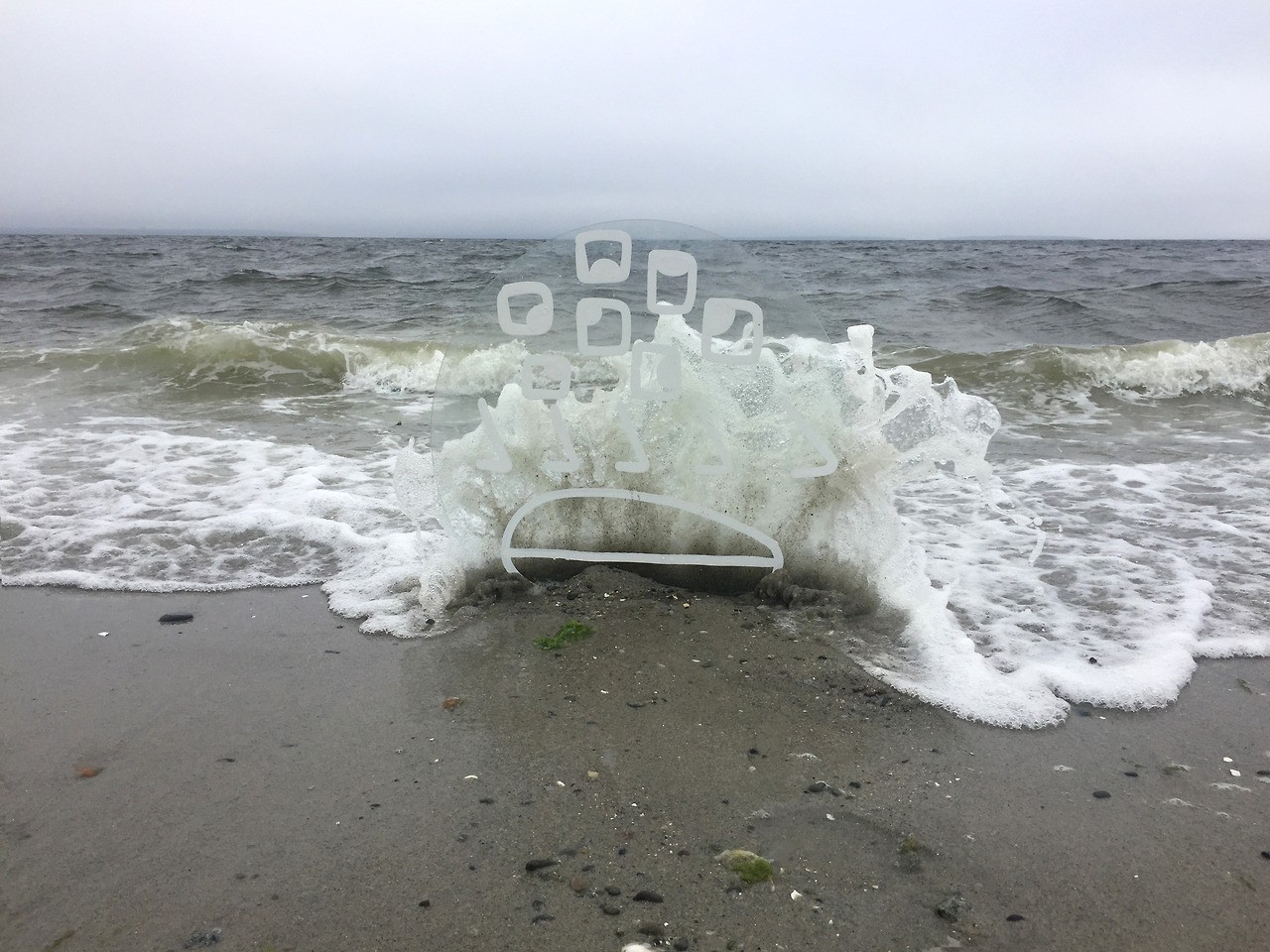 Drawing of a face with seven eyes, five noses and one mouth painted in white on circle piece of glass stuck in sand against wave. 